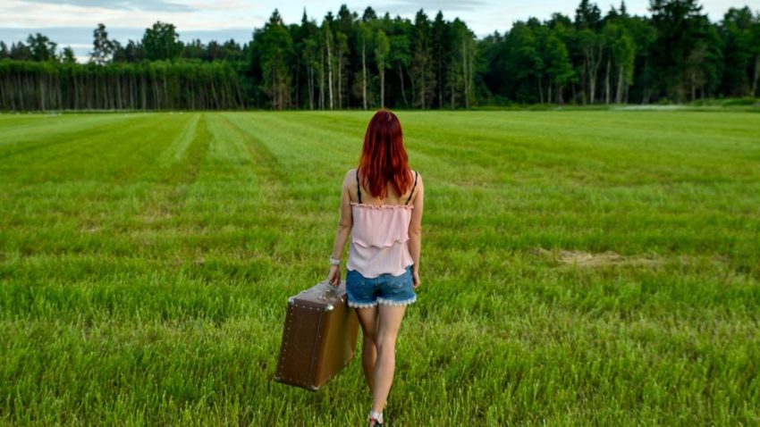 Woman with red hair and vintage suitcase walking through Kolga's lush meadow in Estonia on a summer day.
